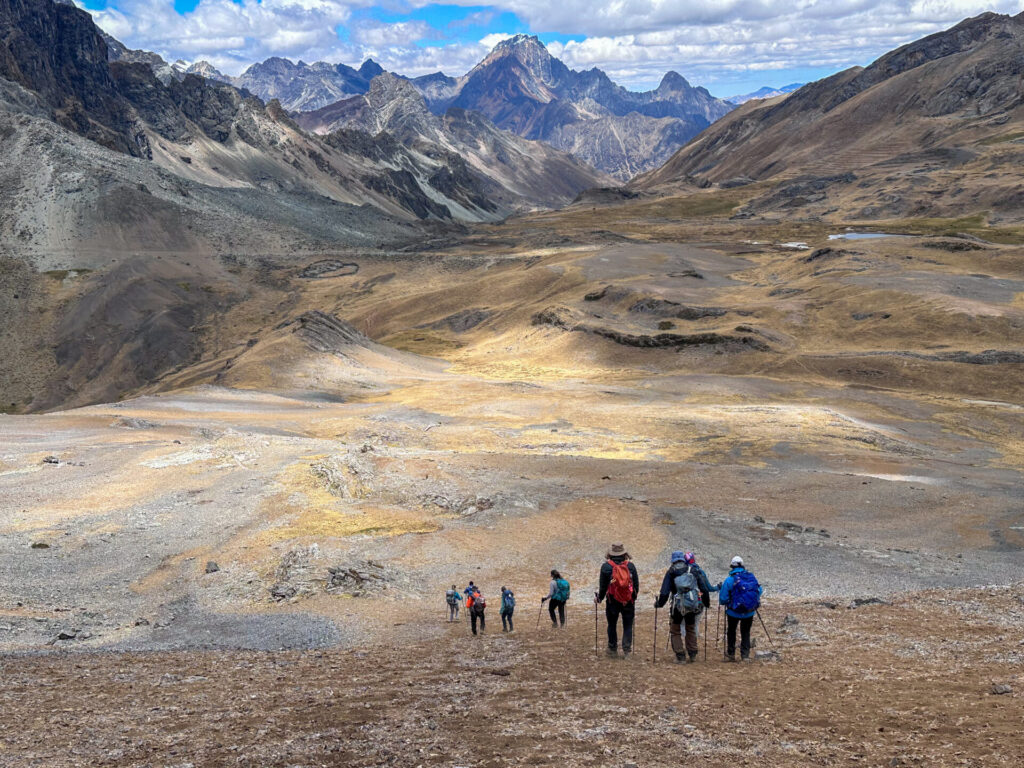 Des randonneurs munis de sacs à dos descendent un versant rocheux et aride de la montagne sur le célèbre Trek du Huayhuash, en direction d'une vallée large et ouverte entourée de pics escarpés et de nuages dans le lointain. - Karavaniers