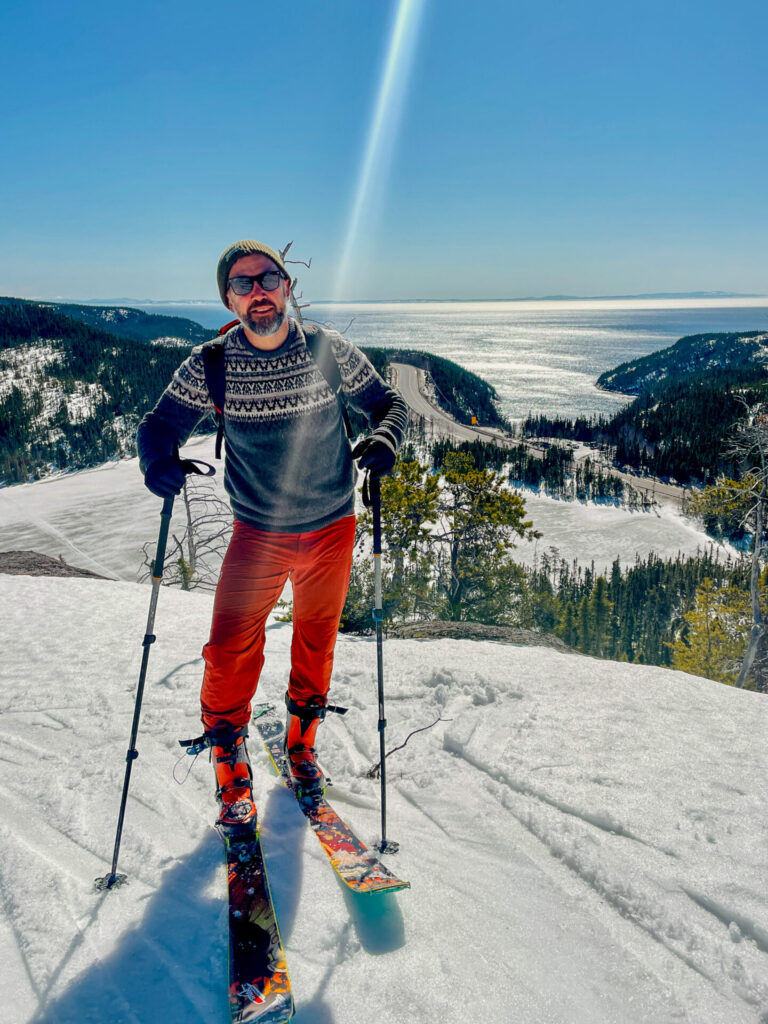 Une personne portant un pull à motifs, un pantalon orange et des lunettes de soleil se tient debout sur des skis avec des bâtons sur une colline enneigée dans les montagnes d'Uapishka, surplombant les arbres, l'eau et les sommets des monts Groulx sous un ciel bleu clair. - Karavaniers