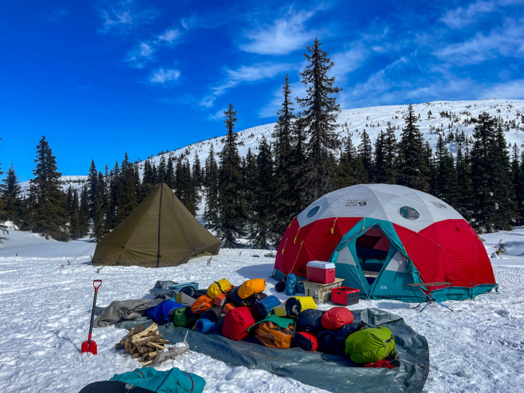 Deux tentes sont installées dans la neige de l'Uapishka (monts Groulx), entourées de pins. Du matériel de plein air et de ski, ainsi que des sacs à dos colorés, sont éparpillés sur une bâche. Une pelle et du bois de chauffage reposent sous un ciel bleu parsemé de nuages. - Karavaniers