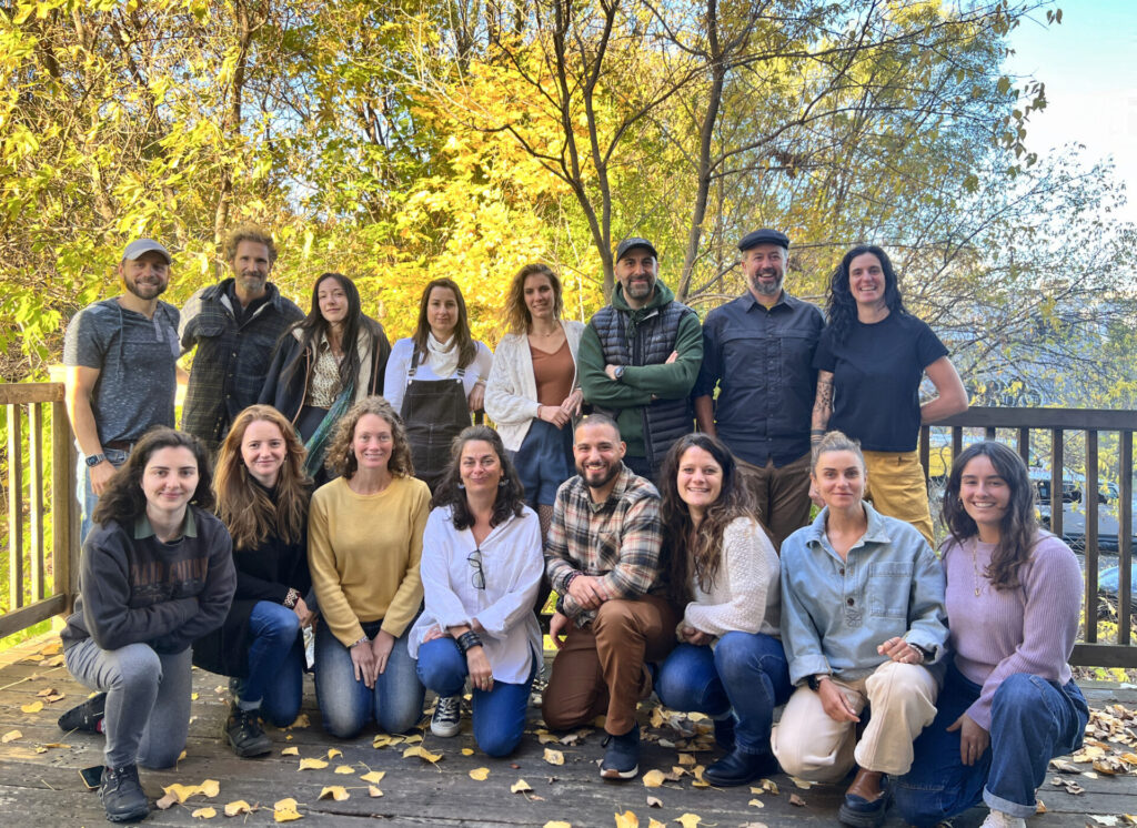 Un groupe de seize adultes pose pour une photo sur une terrasse en bois entourée d'arbres d'automne. Certains sont debout, d'autres sont assis ou agenouillés devant. Des feuilles jaunes recouvrent certaines parties de la terrasse. - Karavaniers
