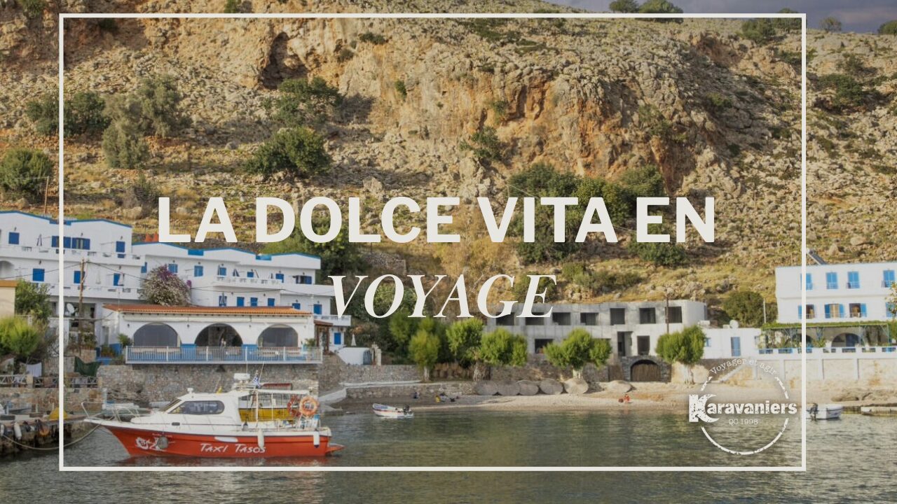 Village côtier avec des bâtiments blancs et une colline rocheuse en arrière-plan, un bateau rouge et blanc sur l'eau au premier plan, et le texte LA DOLCE VITA EN VOYAGE - une scène parfaite pour inspirer votre prochaine conférence ou votre prochain voyage. - Karavaniers