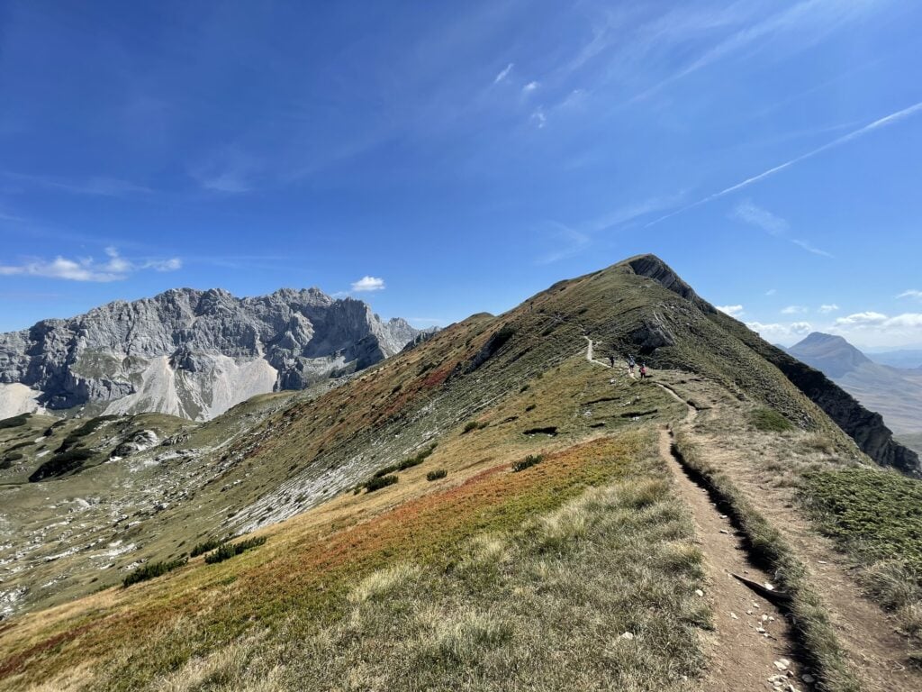 Un étroit sentier de terre serpente le long d'une crête montagneuse herbeuse dans les Alpes Dinariques sous un ciel bleu, avec des pics rocheux en arrière-plan et quelques randonneurs visibles au loin. - Karavaniers