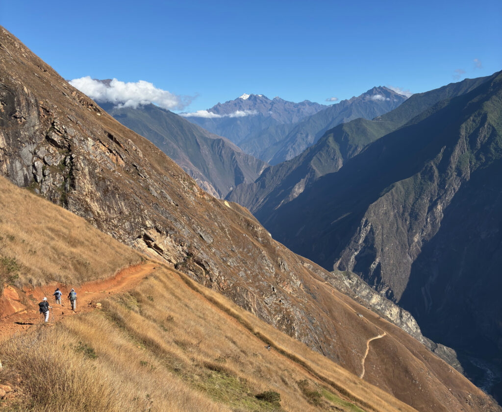 Trois randonneurs marchent sur un étroit sentier de terre battue sur un versant de montagne herbeux et escarpé - une scène inspirante pour un voyage sur mesure au Pérou - avec des montagnes escarpées et des vallées profondes qui s'étendent sous un ciel bleu limpide. - Karavaniers