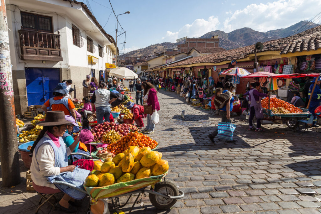 Dans une rue pavée, un marché en plein air très animé propose des fruits et des légumes frais, tandis que les gens font leurs achats sous un ciel ensoleillé - une scène authentique parfaite pour un voyage sur mesure au Pérou, avec des bâtiments traditionnels et des montagnes en arrière-plan. - Karavaniers