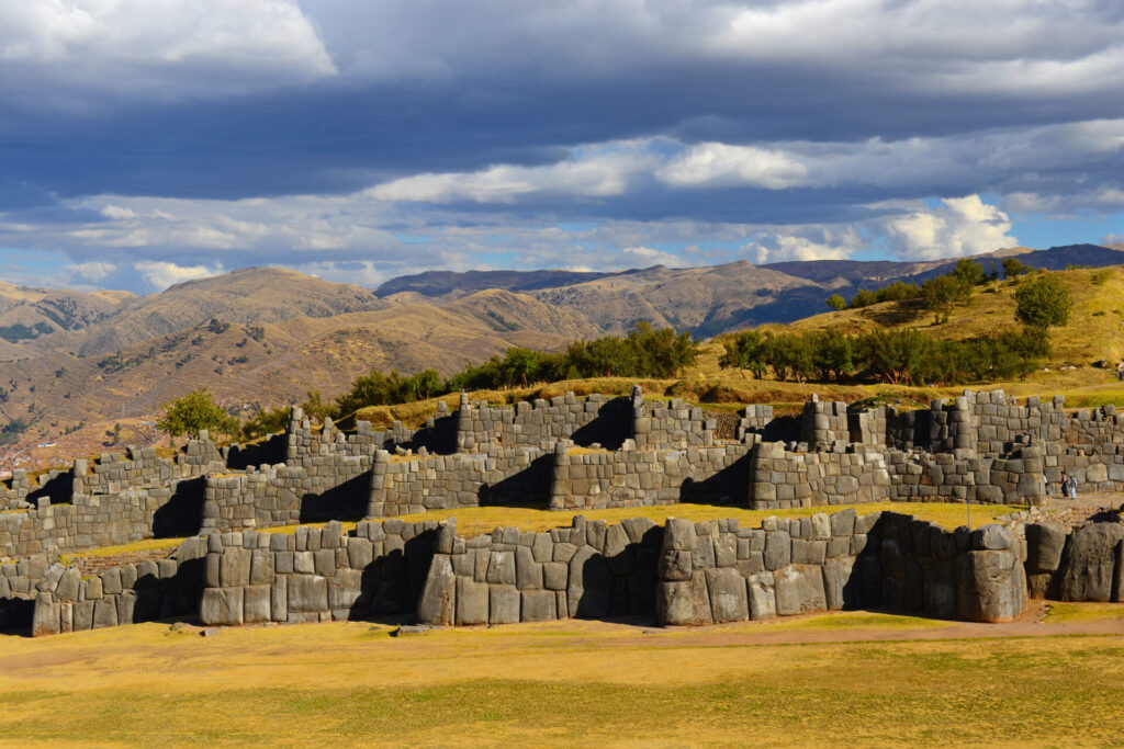 Explorez les ruines de Sacsayhuamán au Pérou, où de grandes pierres taillées avec précision forment de superbes terrasses - un moment inoubliable de tout voyage sur mesure au Pérou au milieu de terrains herbeux et de montagnes majestueuses sous un ciel nuageux. - Karavaniers