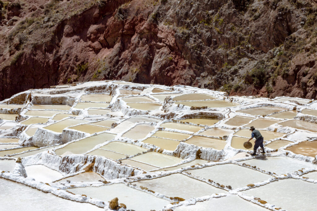 Une personne traverse des bassins géométriques d'évaporation du sel construits à flanc de colline - une scène emblématique à découvrir lors d'un voyage sur mesure au Pérou - entourés d'un terrain rocheux brun et de bassins peu profonds séparés par des barrières blanches incrustées de sel. - Karavaniers