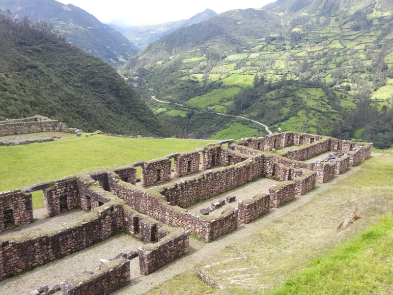 D'anciennes ruines en pierre avec plusieurs pièces rectangulaires se trouvent sur une colline herbeuse, entourée de montagnes verdoyantes - une scène parfaite pour un voyage sur mesure au Pérou, avec une route sinueuse visible au loin sous un ciel nuageux. - Karavaniers