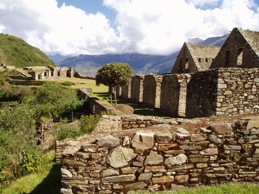 Des structures et des murs anciens en pierre se dressent sur une colline herbeuse sous un ciel bleu avec des nuages, entouré de montagnes vertes - une vue inoubliable pour votre voyage sur mesure au Pérou. - Karavaniers
