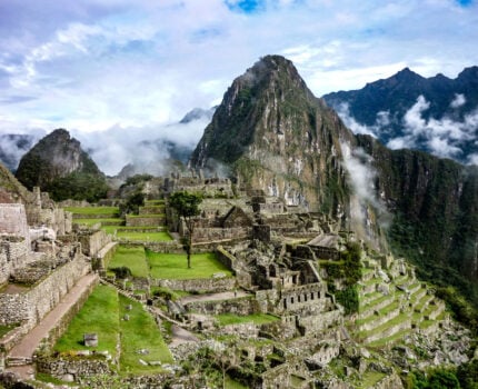 Les anciennes ruines de pierre du Machu Picchu sont situées sur une crête montagneuse verdoyante, où les randonneurs parcourent le Camino Inca au Pérou au milieu de grands pics et de nuages sous un ciel partiellement nuageux. - Karavaniers