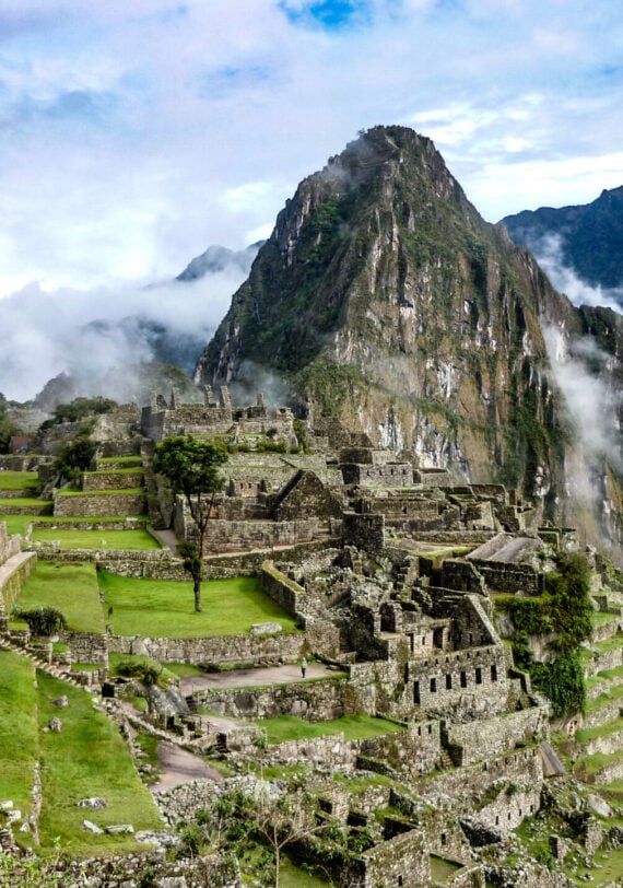 Les anciennes ruines de pierre du Machu Picchu sont situées sur une crête montagneuse verdoyante, où les randonneurs parcourent le Camino Inca au Pérou au milieu de grands pics et de nuages sous un ciel partiellement nuageux. - Karavaniers