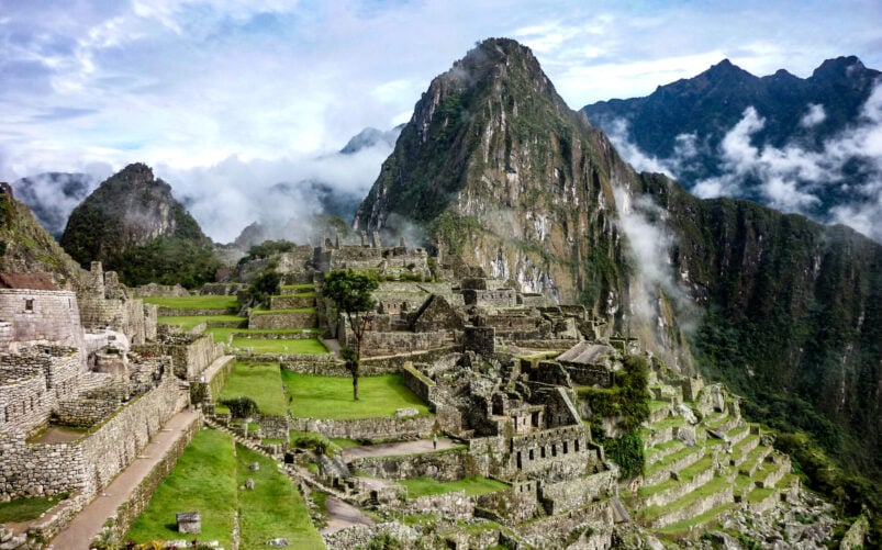 Les anciennes ruines de pierre du Machu Picchu sont situées sur une crête montagneuse verdoyante, où les randonneurs parcourent le Camino Inca au Pérou au milieu de grands pics et de nuages sous un ciel partiellement nuageux. - Karavaniers