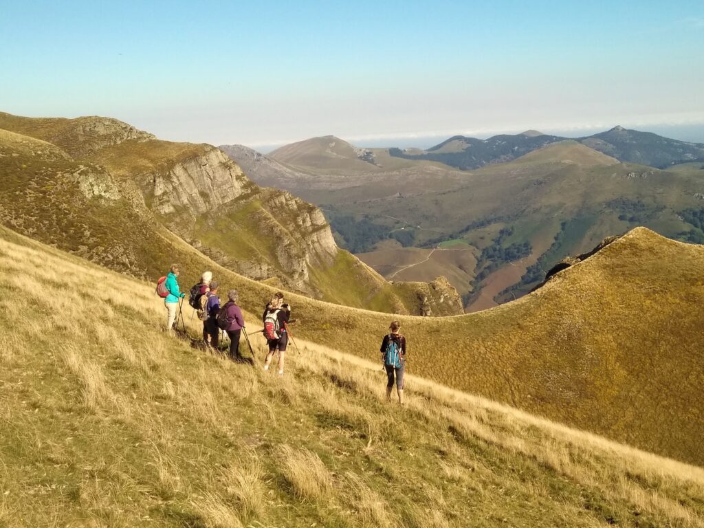 Un groupe de randonneurs marche en file indienne le long d'une pente montagneuse herbeuse avec des crêtes rocheuses et des collines lointaines sous un ciel bleu clair, profitant d'un trek dans les pays basques. - Karavaniers