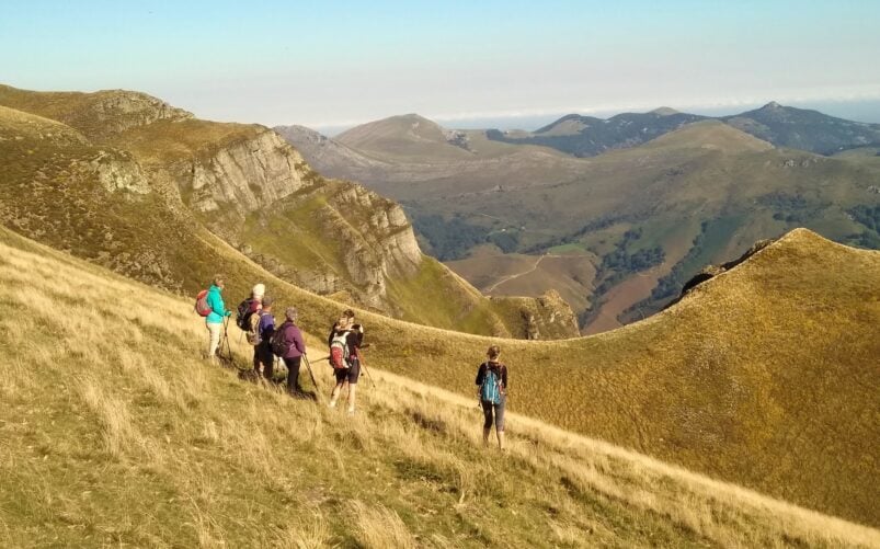 Un groupe de randonneurs marche en file indienne le long d'une pente montagneuse herbeuse avec des crêtes rocheuses et des collines lointaines sous un ciel bleu clair, profitant d'un trek dans les pays basques. - Karavaniers