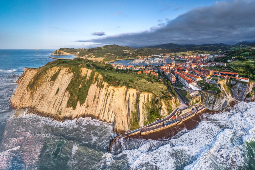 Vue aérienne des falaises escarpées du Pays basque, parfaites pour une randonnée dans les pays basques ; ville balnéaire aux toits rouges, routes sinueuses, collines verdoyantes et vagues déferlantes sous un ciel partiellement nuageux. - Karavaniers