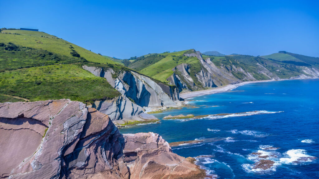La côte rocheuse, avec ses collines verdoyantes et abruptes, ses falaises déchiquetées et ses couches de formations rocheuses exposées, rencontre l'océan bleu sous un ciel clair - un cadre idéal pour un Trek dans les pays basques, alors que les vagues s'écrasent le long du paysage accidenté. - Karavaniers