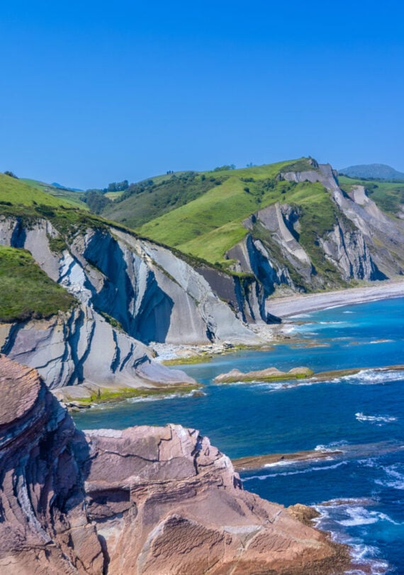 La côte rocheuse, avec ses collines verdoyantes et abruptes, ses falaises déchiquetées et ses couches de formations rocheuses exposées, rencontre l'océan bleu sous un ciel clair - un cadre idéal pour un Trek dans les pays basques, alors que les vagues s'écrasent le long du paysage accidenté. - Karavaniers