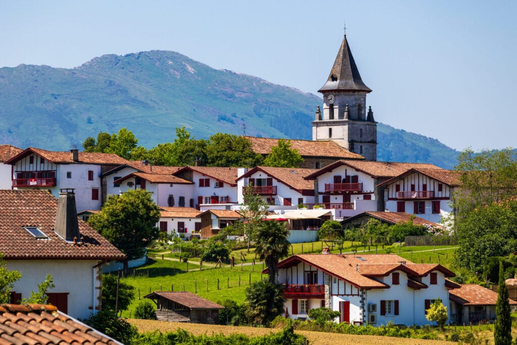 Un village aux bâtiments blancs et aux toits rouges se dresse dans un paysage verdoyant, idéal pour une randonnée dans les pays basques, avec un clocher d'église s'élevant au-dessus des maisons et une montagne en arrière-plan sous un ciel bleu clair. - Karavaniers