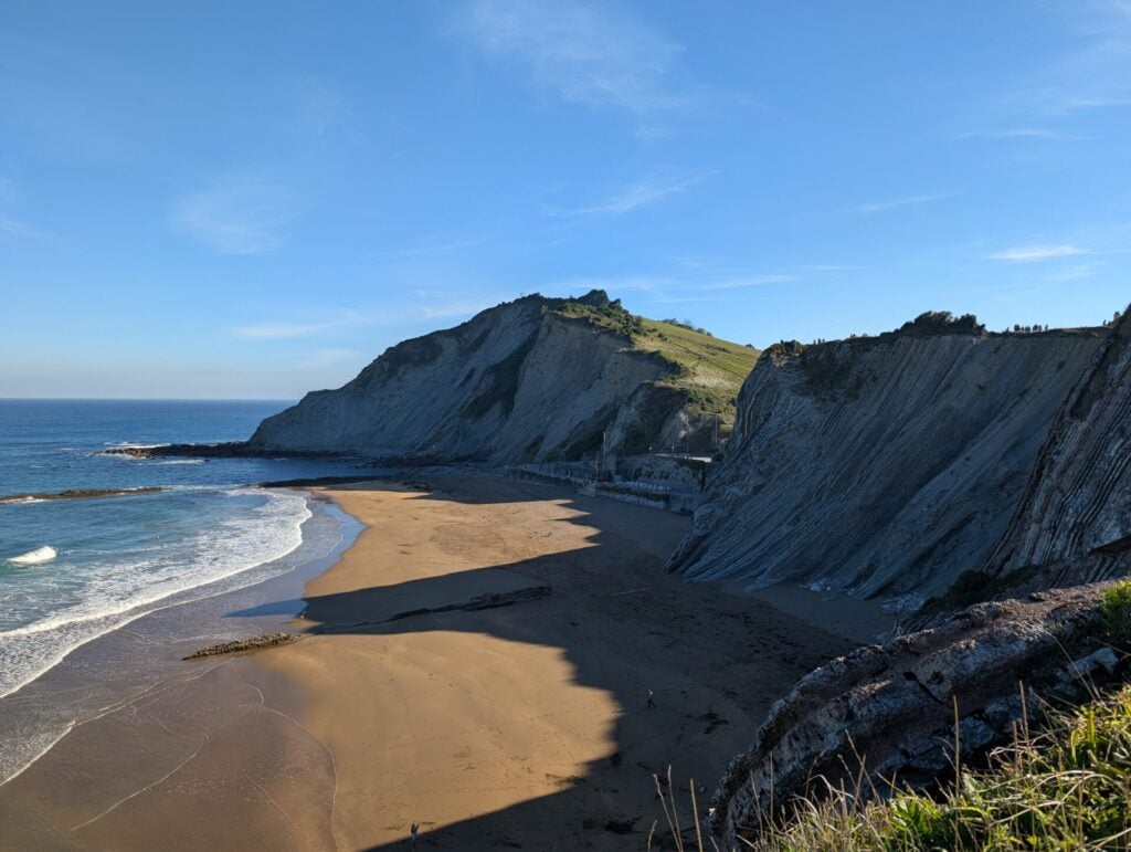 Une plage de sable bordée de falaises rocheuses abruptes rencontre l'océan bleu sous un ciel clair. Parfaites pour une randonnée dans les pays basques, les falaises projettent des ombres sombres, tandis que la végétation verte couronne les hauteurs en arrière-plan. - Karavaniers