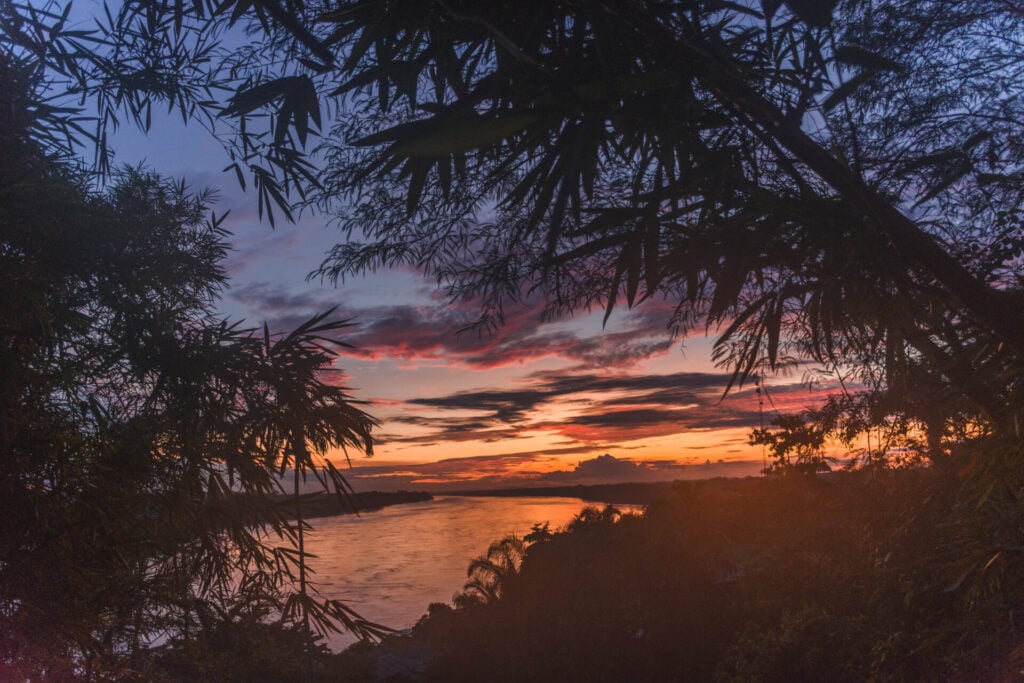 Une rivière au coucher du soleil est partiellement encadrée par des silhouettes de branches et de feuilles, évoquant l'esprit d'un voyage au Pérou. Le ciel est parsemé de nuages orange, jaunes et violets qui se reflètent dans l'eau, tandis qu'un feuillage dense et des arbres bordent la rive. - Karavaniers