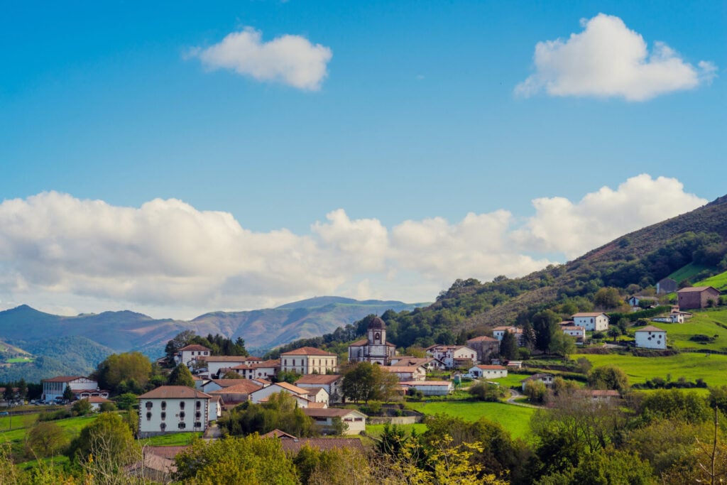 Un petit village aux bâtiments blancs et aux toits rouges se trouve au milieu de collines verdoyantes, sous un ciel bleu parsemé de nuages. Un décor idyllique parfait pour un Trek dans les pays basques, avec des montagnes boisées en arrière-plan. - Karavaniers