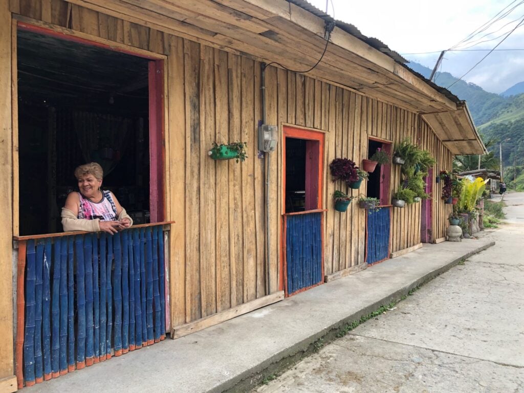 Une maison en bois avec des fenêtres bordées de rouge et des panneaux inférieurs bleus. Des plantes en pot sont exposées sur le mur. Une femme âgée sourit dans l'embrasure de la porte et des montagnes sont visibles à l'arrière-plan. - Karavaniers