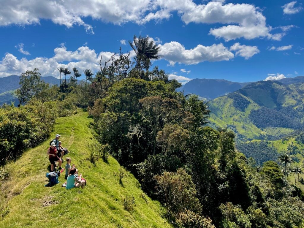 Un groupe de personnes est assis ou debout sur une colline herbeuse surplombant une vallée verdoyante avec de hautes montagnes et des arbres sous un ciel bleu vif avec des nuages blancs épars. - Karavaniers
