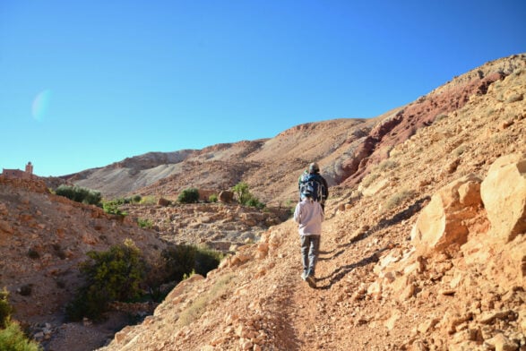 Deux personnes marchent sur un chemin rocailleux en montée dans un paysage désertique avec des collines brunes sous un ciel bleu clair pendant leur voyage au Monténégro. On aperçoit à l'arrière-plan une végétation verte clairsemée et un petit bâtiment. - Karavaniers