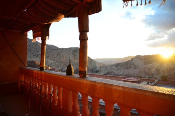 Un balcon couvert avec des colonnes décoratives donne sur les collines et les montagnes escarpées éclairées par le soleil au coucher du soleil - une scène parfaite d'un Voyage au Monténégro, où la lumière dorée baigne le paysage sous un ciel partiellement nuageux. - Karavaniers