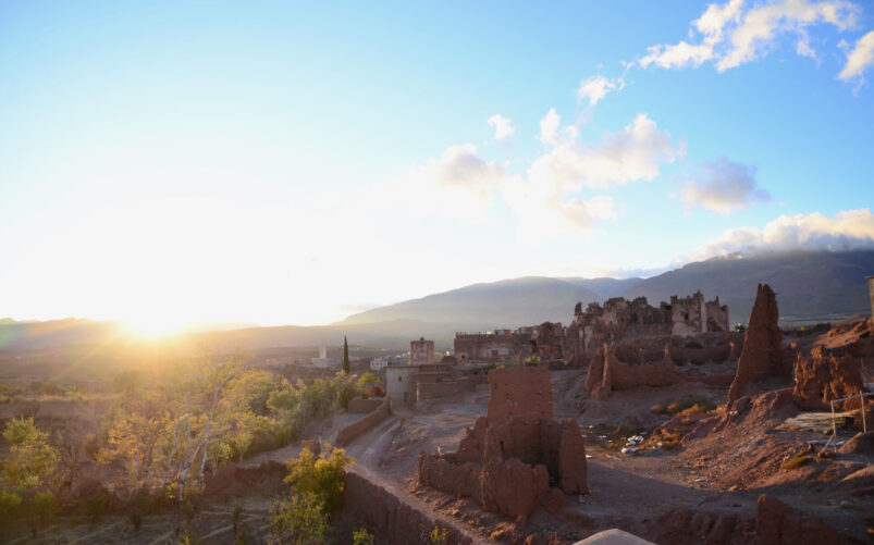 L'image évoque un Voyage au Monténégro, avec les ruines de vieux bâtiments en pisé dans un paysage désertique au coucher du soleil. Des montagnes et des nuages encadrent l'arrière-plan, tandis que la lumière du soleil provenant de la gauche éclaire partiellement cette scène évocatrice. - Karavaniers