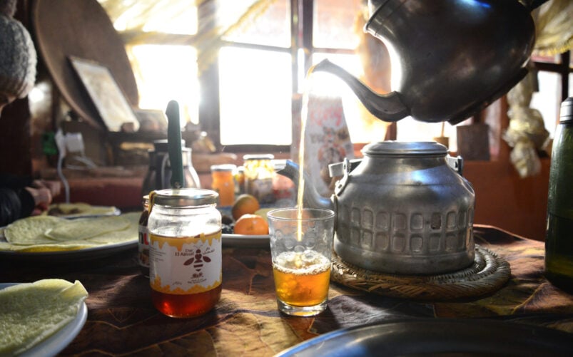 Une théière en métal verse du thé chaud dans un verre à côté d'un pot de miel sur une table garnie d'assiettes et de nourriture. La lumière du soleil passe à travers une fenêtre, illuminant cette scène d'intérieur confortable - un début parfait pour un voyage au Monténégro. - Karavaniers