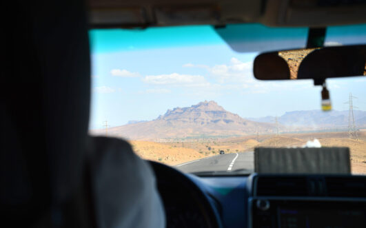 Vue de l'intérieur d'une voiture lors d'un voyage au Monténégro, sur une route pavée traversant un paysage désertique avec des montagnes lointaines sous un ciel bleu. Au premier plan, on aperçoit une partie du conducteur et de l'intérieur de la voiture. - Karavaniers