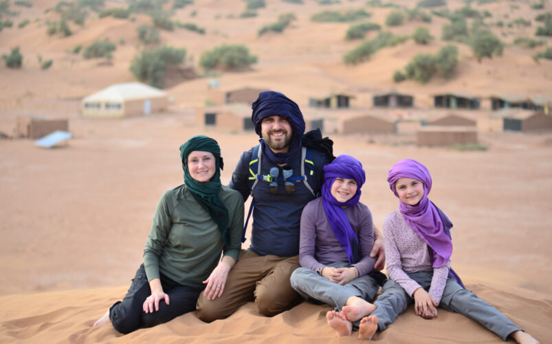 Quatre personnes, deux adultes et deux enfants, sont assises sur une dune de sable, portant foulard et vêtements décontractés. Les tentes et la verdure éparpillée à l'arrière-plan évoquent l'esprit d'aventure - un véritable voyage au Monténégro dans un cadre désertique. - Karavaniers