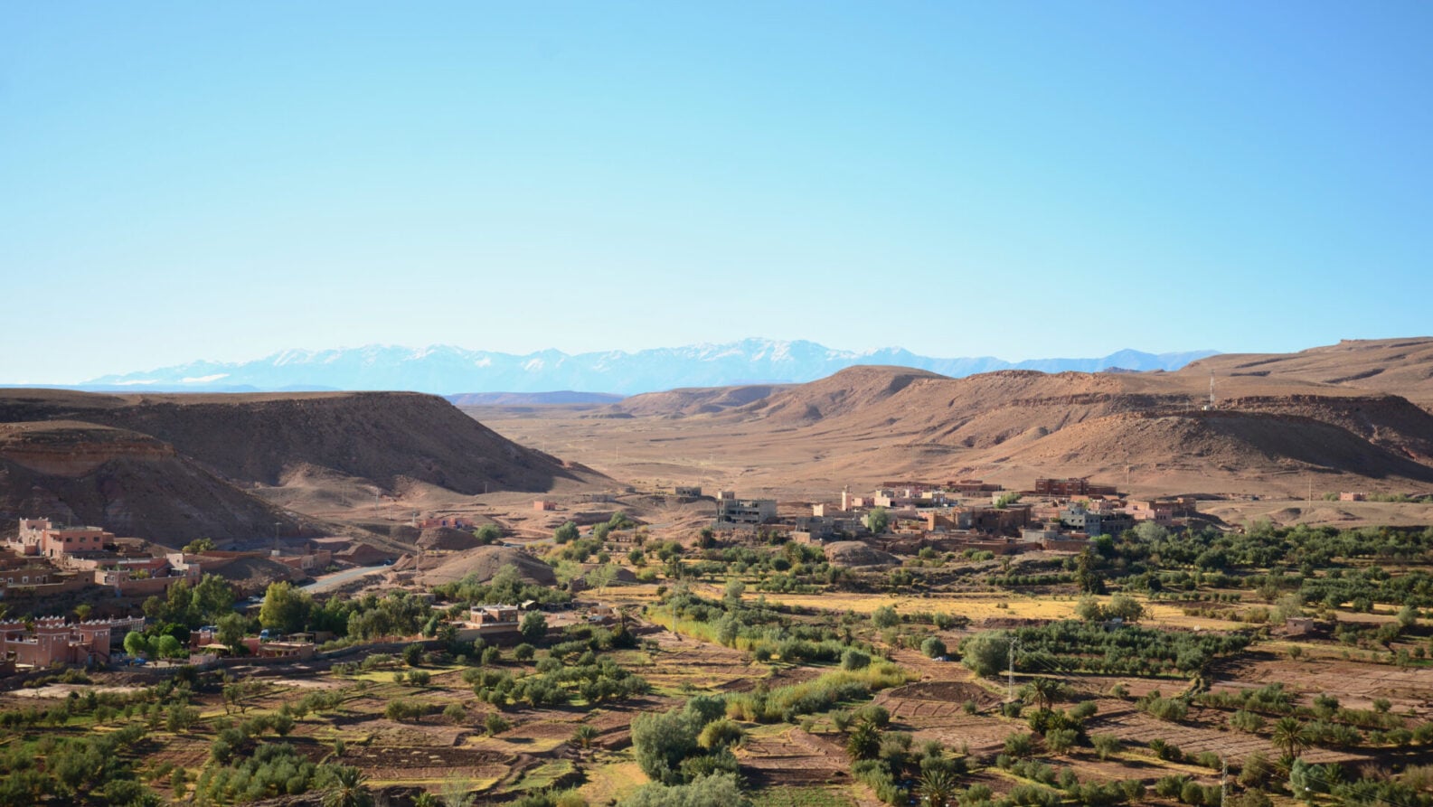 Une vue large d'un paysage rural avec des groupes de terres agricoles vertes et d'arbres au premier plan, un petit village aux bâtiments rougeâtres et des collines arides - parfaites pour un voyage au Monténégro - entourées de montagnes enneigées sous un ciel bleu limpide. - Karavaniers