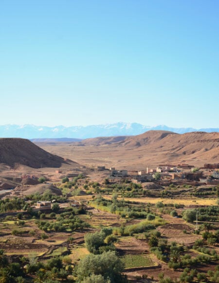 Une vue large d'un paysage rural avec des groupes de terres agricoles vertes et d'arbres au premier plan, un petit village aux bâtiments rougeâtres et des collines arides - parfaites pour un voyage au Monténégro - entourées de montagnes enneigées sous un ciel bleu limpide. - Karavaniers