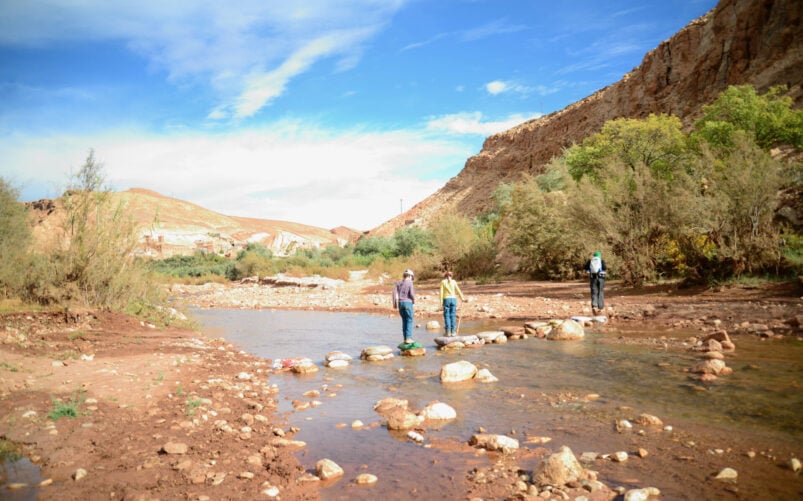 Trois personnes traversent un ruisseau peu profond sur de gros rochers dans un paysage aride de collines et de buissons, sous un ciel partiellement nuageux. Une quatrième personne se tient à l'écart au bord de l'eau - une scène aventureuse d'un Voyage au Monténégro. - Karavaniers