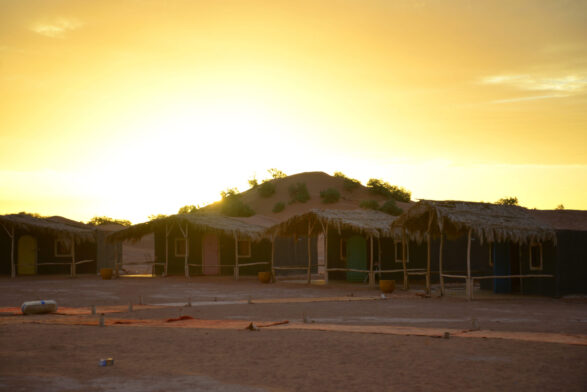 Une rangée de huttes aux toits de chaume se dresse sur un sol sablonneux, avec une colline et une végétation clairsemée en arrière-plan. Le soleil se couche derrière la colline, jetant une chaude lueur jaune sur cette scène paisible d'un voyage au Monténégro. - Karavaniers
