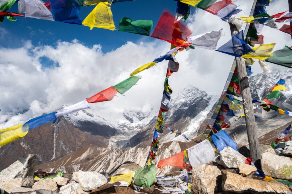Des drapeaux de prière colorés sont suspendus à des poteaux en bois sur un sommet rocheux, avec des montagnes enneigées et des nuages visibles à l'arrière-plan dans un ciel bleu. - Karavaniers