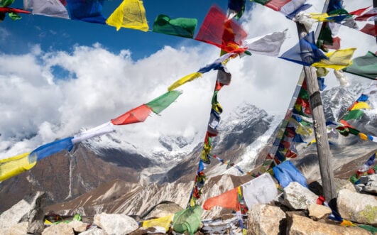Des drapeaux de prière colorés sont suspendus à des poteaux en bois sur un sommet rocheux, avec des montagnes enneigées et des nuages visibles à l'arrière-plan dans un ciel bleu. - Karavaniers