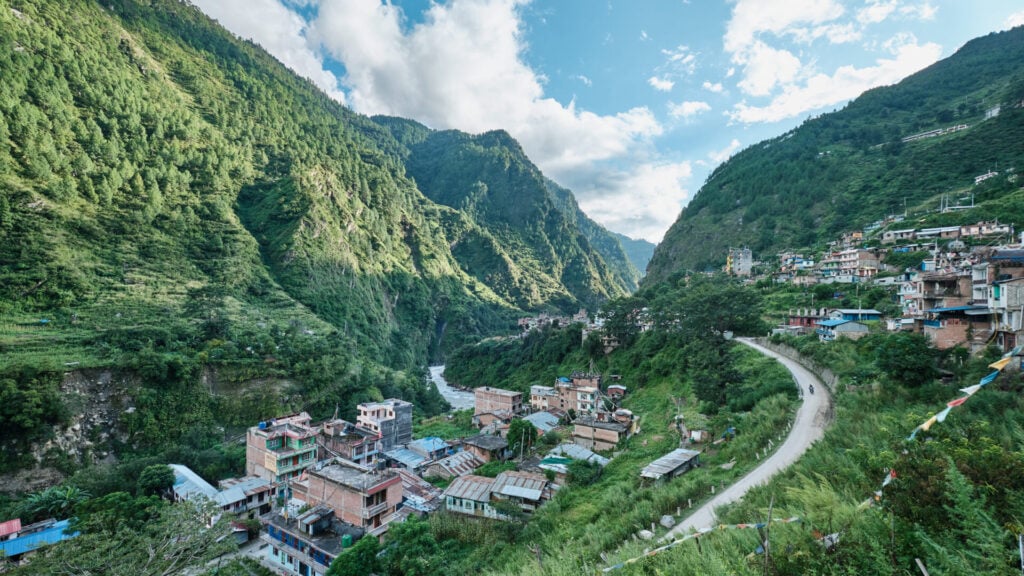 Une petite ville aux bâtiments colorés se trouve dans une vallée montagneuse verdoyante. Des collines verdoyantes et boisées s'élèvent de part et d'autre, et une route sinueuse traverse la scène sous un ciel partiellement nuageux. - Karavaniers