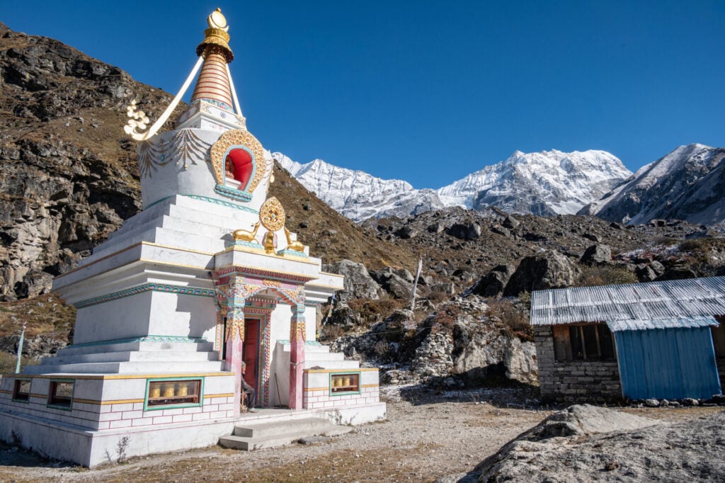 Un stupa blanc aux décorations colorées se dresse sur un terrain rocailleux près d'un bâtiment au toit bleu, avec des montagnes enneigées visibles à l'arrière-plan sous un ciel bleu clair. - Karavaniers