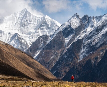Une personne vêtue d'une veste rouge se tient sur l'herbe dans une vallée, entourée de grandes montagnes escarpées aux sommets enneigés, sous un ciel partiellement nuageux. - Karavaniers