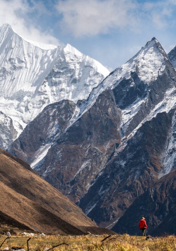 Une personne vêtue d'une veste rouge se tient sur l'herbe dans une vallée, entourée de grandes montagnes escarpées aux sommets enneigés, sous un ciel partiellement nuageux. - Karavaniers
