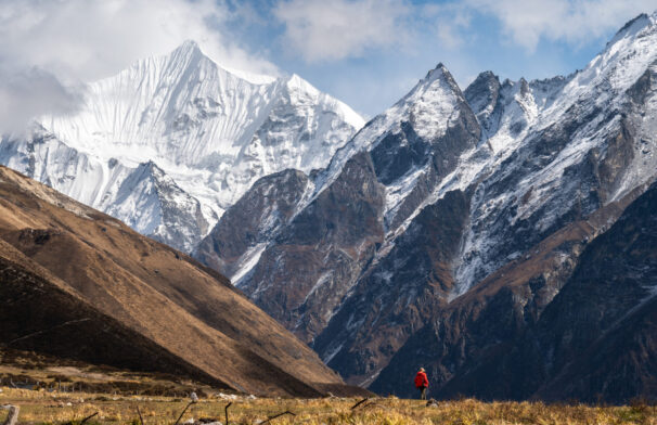 Une personne vêtue d'une veste rouge se tient sur l'herbe dans une vallée, entourée de grandes montagnes escarpées aux sommets enneigés, sous un ciel partiellement nuageux. - Karavaniers