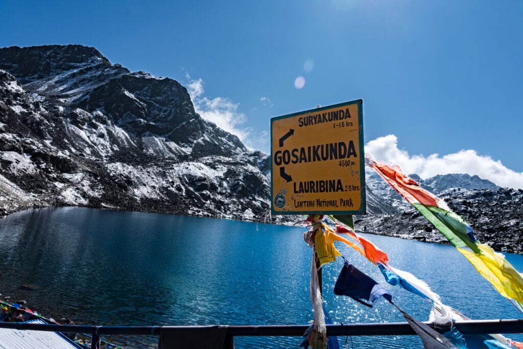Un panneau se dresse au bord d'un lac de montagne d'un bleu limpide, indiquant les directions de Suryakunda, Gosaikunda et Lauribina. Des drapeaux de prière colorés sont attachés au poteau, avec en arrière-plan des pics enneigés et des pentes rocheuses. - Karavaniers