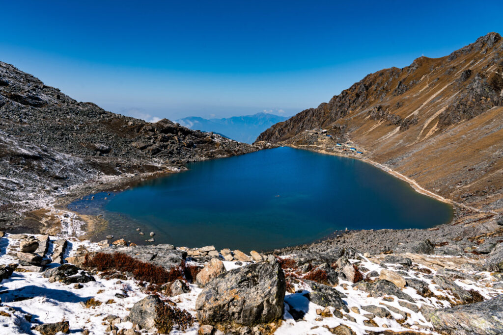Un lac bleu clair entouré de montagnes rocheuses et enneigées sous un ciel bleu vif. Le paysage présente une végétation clairsemée et des collines lointaines sont visibles à l'arrière-plan. - Karavaniers