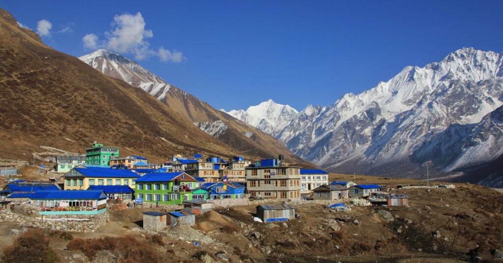 Un petit village aux bâtiments à toits bleus se trouve au pied de montagnes escarpées et enneigées, sous un ciel bleu clair. Le paysage est rocailleux et sec au premier plan, avec des taches de neige visibles. - Karavaniers