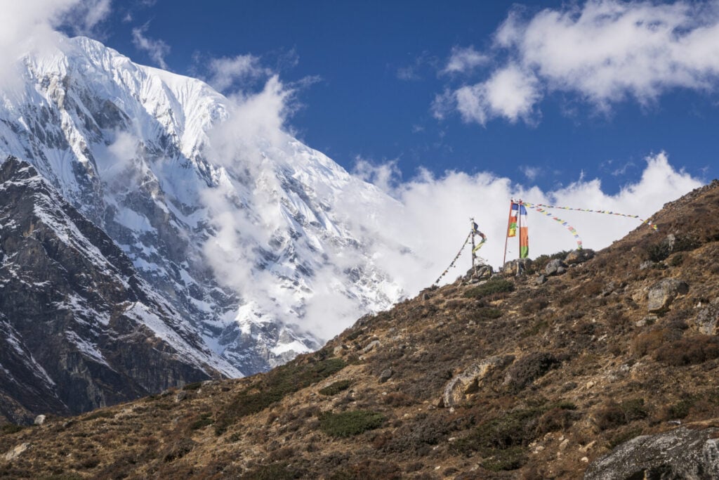 En arrière-plan, une montagne enneigée dont le sommet est partiellement masqué par les nuages. Au premier plan, des drapeaux de prière sont suspendus le long d'une colline rocheuse - une scène typique d'un Trek sur mesure dans la vallée du Langtang au Népal. - Karavaniers