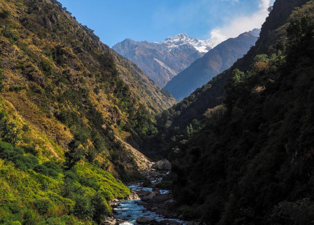 Une rivière étroite coule dans une vallée montagneuse verdoyante, avec des collines hautes et escarpées de part et d'autre et des sommets enneigés visibles à l'arrière-plan sous un ciel bleu limpide. - Karavaniers