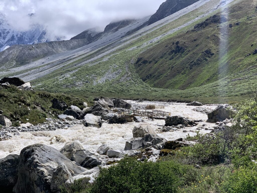 Une rivière au cours rapide traverse un paysage rocheux avec des collines vertes et des montagnes en arrière-plan. Un petit pont en bois traverse la rivière et des nuages couvrent partiellement les sommets. - Karavaniers