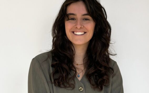 Sarah Haineault, avec ses longs cheveux noirs ondulés et sa chemise grise boutonnée, se tient sur un fond blanc uni, souriante, les bras vaguement croisés. - Karavaniers